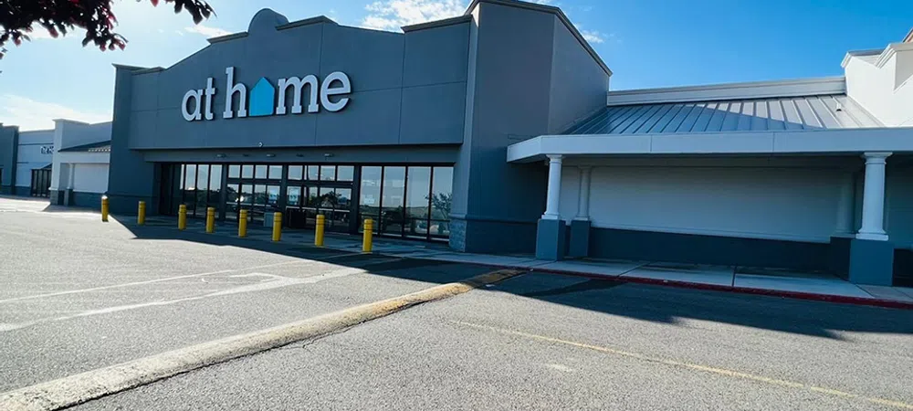 Gray storefront of At Home store with large windows and yellow bollards under a clear blue sky