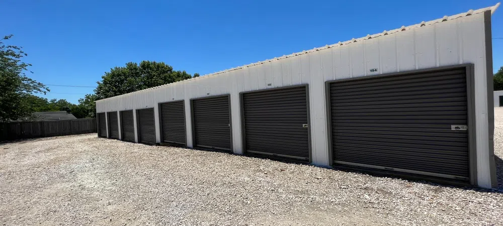 Row of white storage units with closed black roll-up doors under clear blue sky on gravel ground.