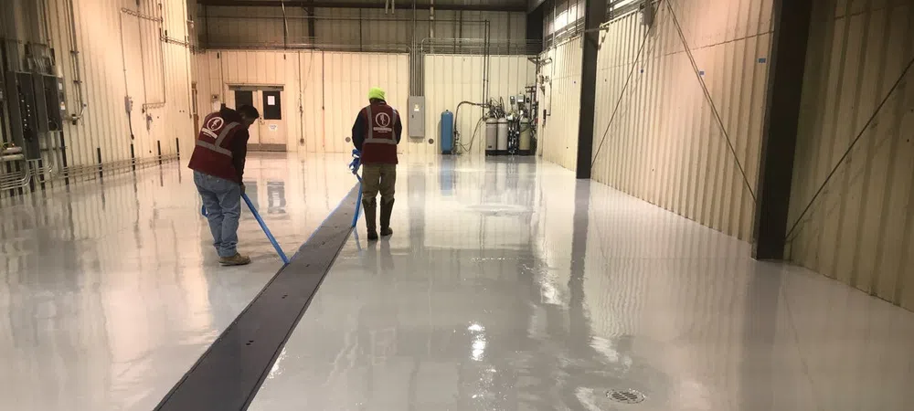 Two workers applying epoxy coating to a clean industrial warehouse floor using rollers under bright lights