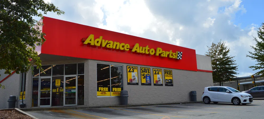 Advance Auto Parts store exterior with red signage, promotional posters, parked cars, and partly cloudy sky.