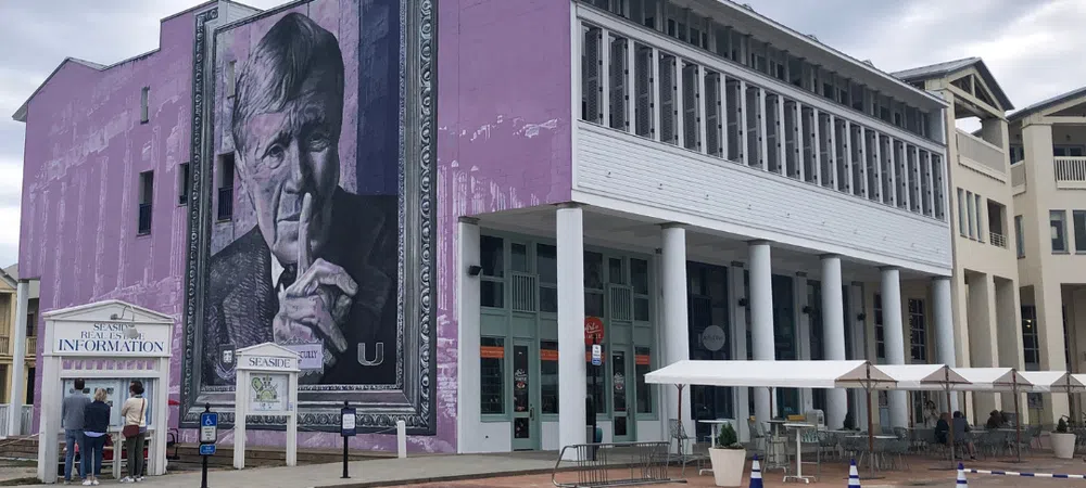 Large purple building with a mural of an elderly man shushing and a tourist information booth outside under cloudy sky.
