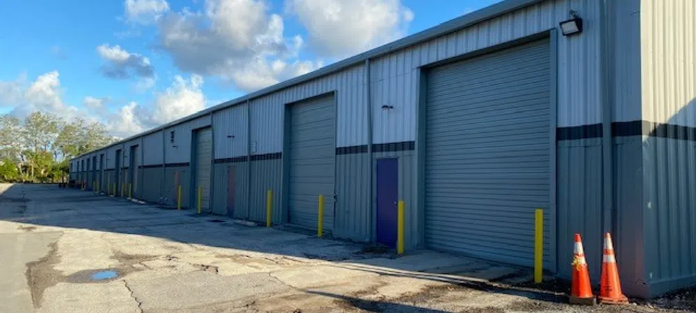 Long metal warehouse building with multiple closed roller doors and yellow bollards under a blue sky.