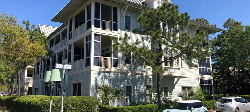 Modern three-story residential building with balconies surrounded by trees and parked cars on a sunny day