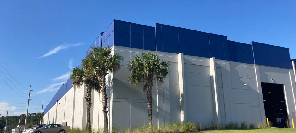 Modern industrial building with blue accents, palm trees, green lawn, and trailer parked outside under clear sky