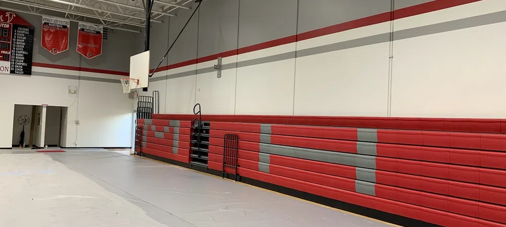 Empty gymnasium with red and gray retractable bleachers and basketball hoop on a polished floor.