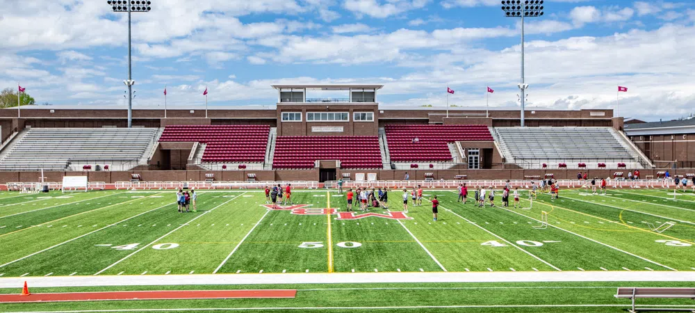 Wide view of a football field with players practicing and empty stadium seating under a partly cloudy sky