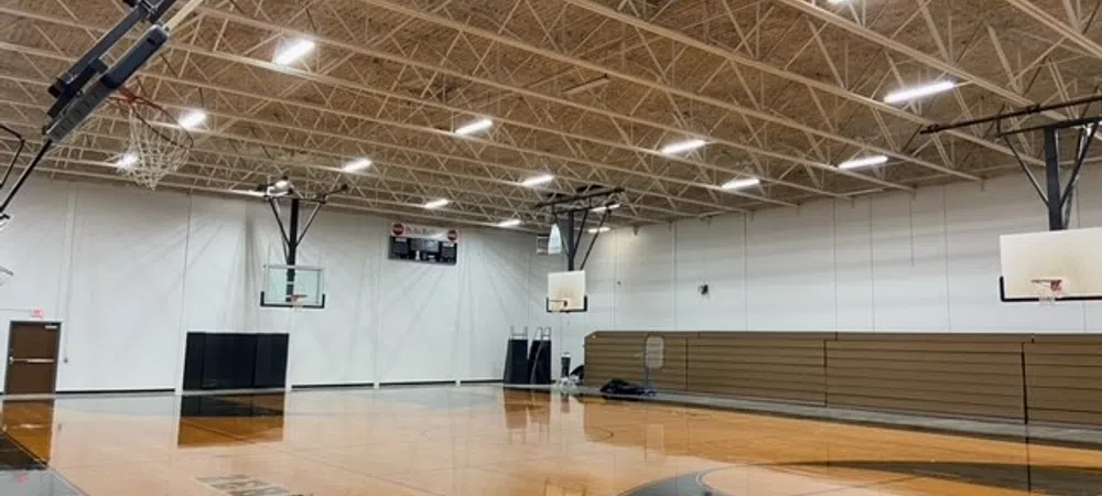 Empty indoor basketball court with wooden floor, multiple hoops, bright ceiling lights, and bleachers on side.