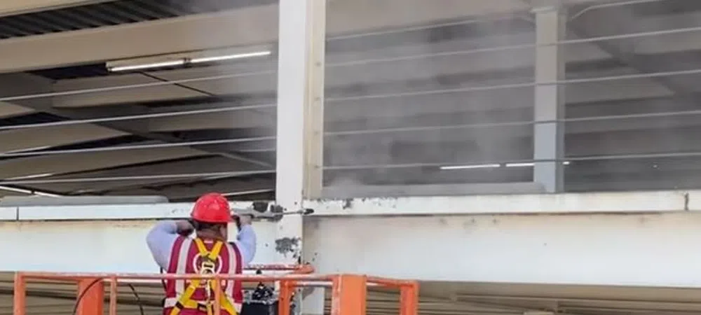 Construction worker in red helmet using a lift to clean or repair a white concrete parking structure beam.