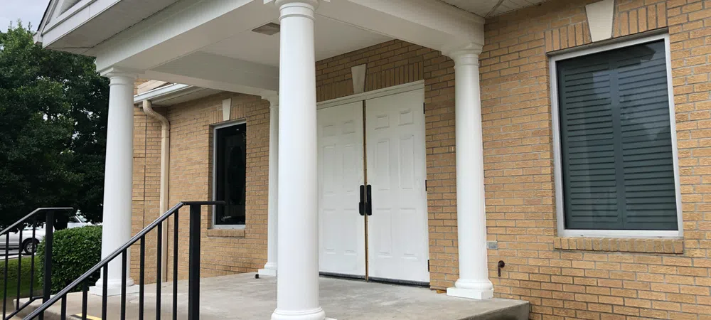 Entrance to a brick building with white double doors, four white columns, and concrete steps with black railing.