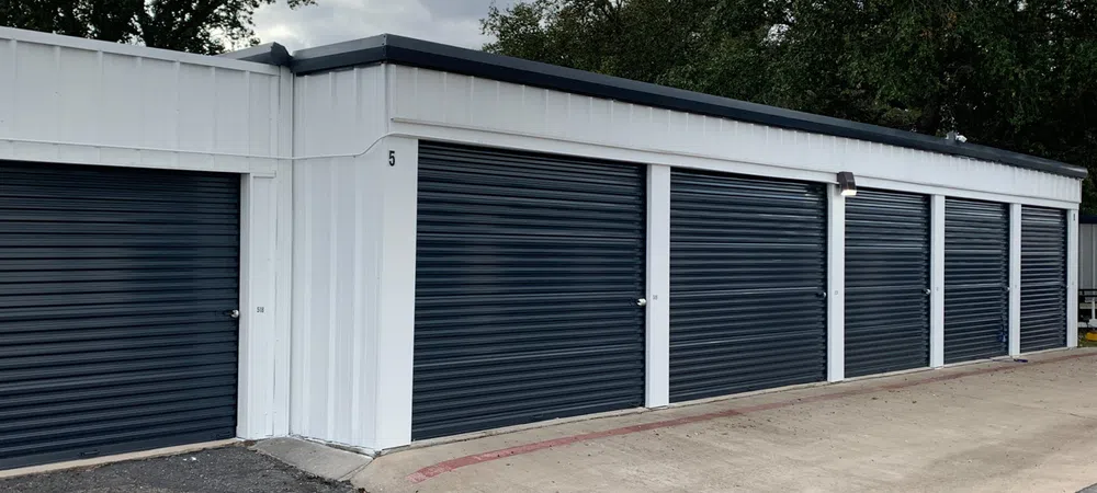 Row of white storage units with dark blue roll-up doors under a cloudy sky and surrounded by trees.