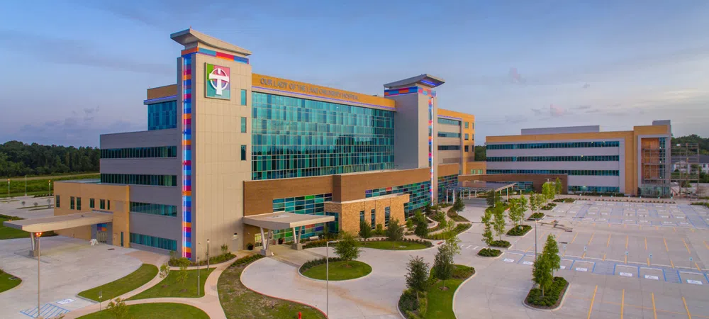 Modern hospital building with glass facade, colorful pillars, surrounding greenery, and spacious parking lot under blue sky.