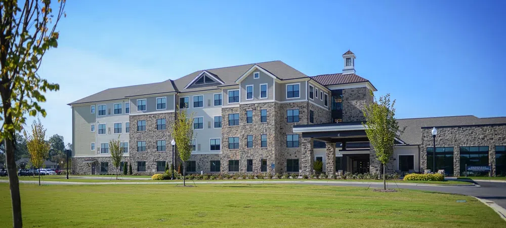 Modern four-story hotel with stone and beige siding exterior under a clear blue sky, surrounded by green lawn and trees.