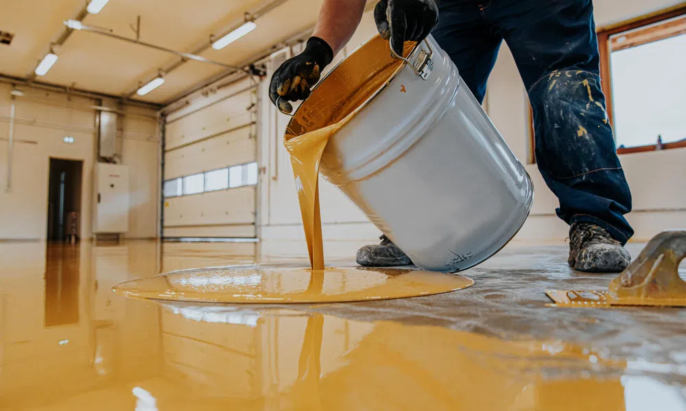 Worker pouring yellow epoxy resin onto floor in industrial space for coating and sealing.