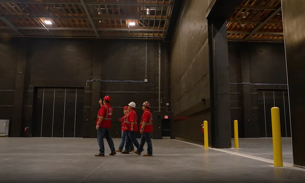 Four construction workers in red safety vests and helmets inspecting a large empty industrial warehouse.