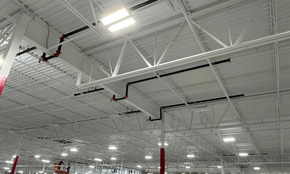 Interior view of a spacious industrial warehouse ceiling with white beams, pipes, and bright LED lights
