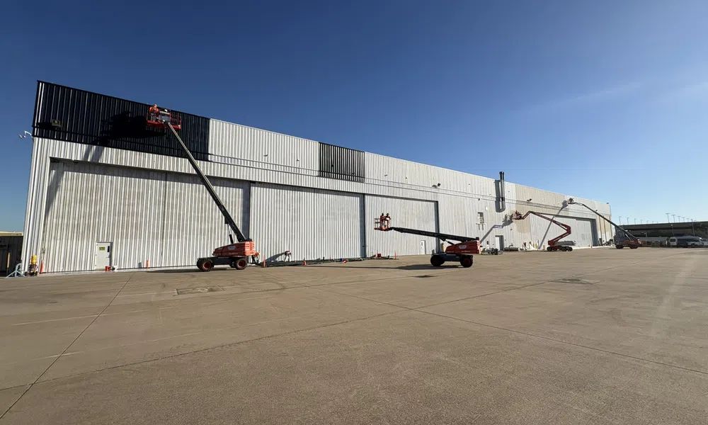 Workers using boom lifts to paint a large industrial warehouse exterior under a clear blue sky.