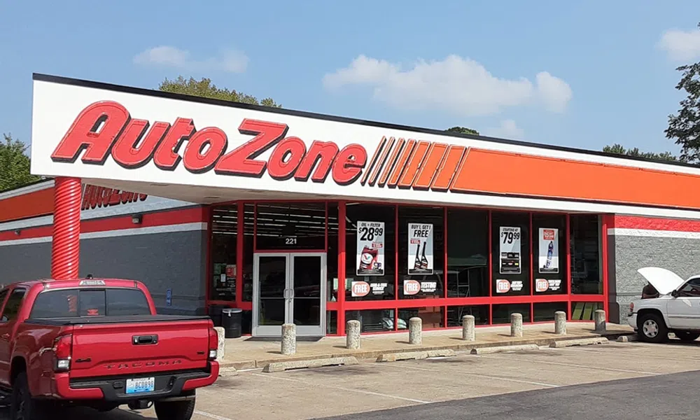 AutoZone storefront with signage, promotional posters in windows, and parked vehicles under a clear sky.