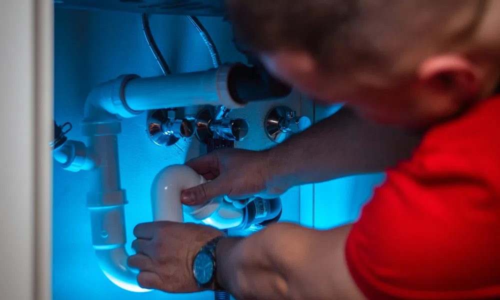 Plumber fixing white PVC pipes under sink illuminated by blue light, wearing red shirt and watch.