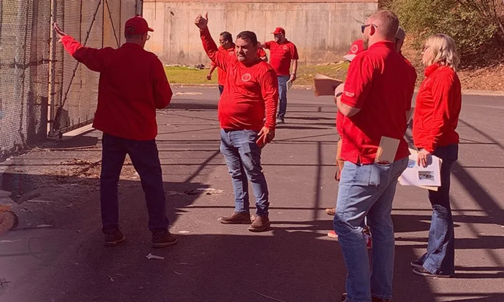 Group of people in red shirts inspecting an outdoor paved area beside a chain-link fence on a sunny day.