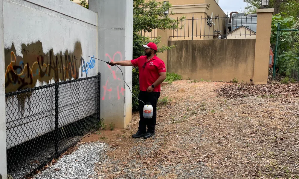 Worker in red uniform spraying graffiti cleaner on concrete wall near building and fence outdoors.