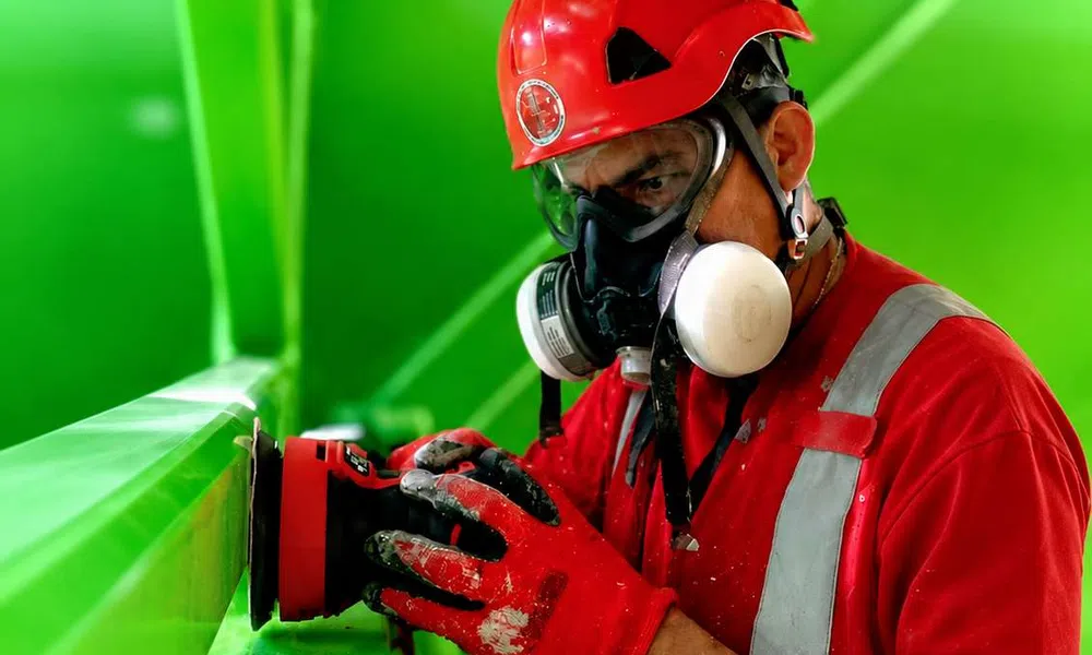 Worker in red gear sanding a bright green metal beam, wearing helmet, gloves, and protective mask indoors.