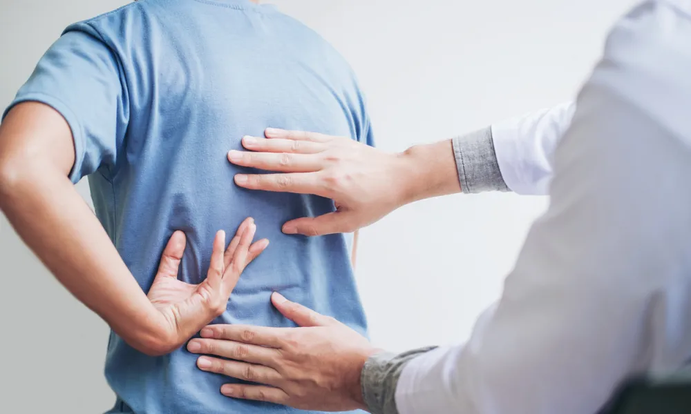 Doctor examining a patient's lower back pain by palpating the spine area in a clinical setting.