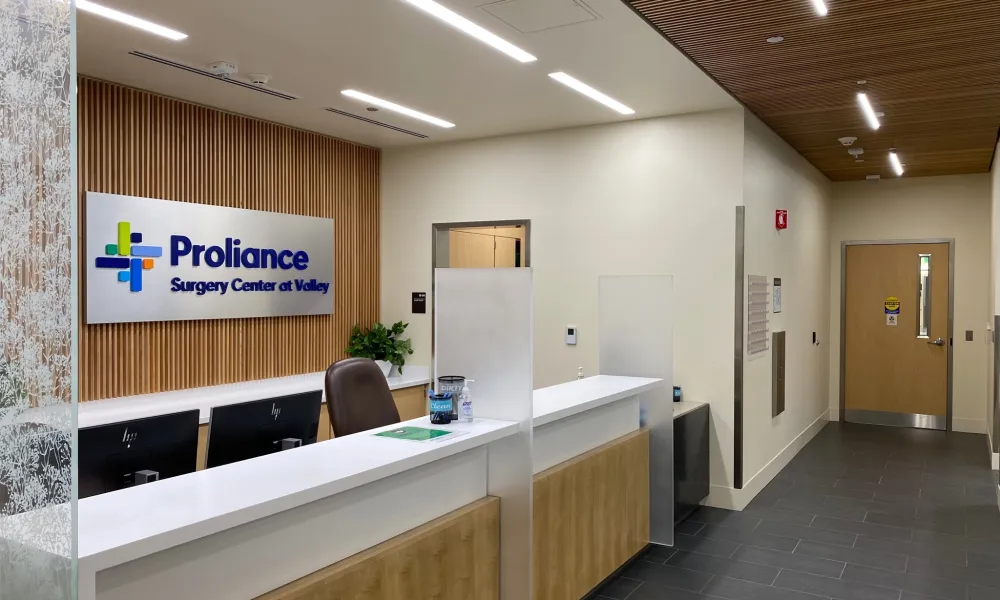 Modern medical reception area with Proliance Surgery Center sign, white desk, wood accents, and hallway.