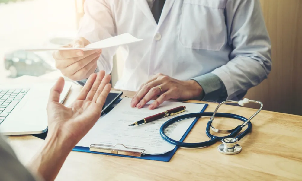 Doctor in white coat handing prescription to patient at desk with stethoscope and clipboard.