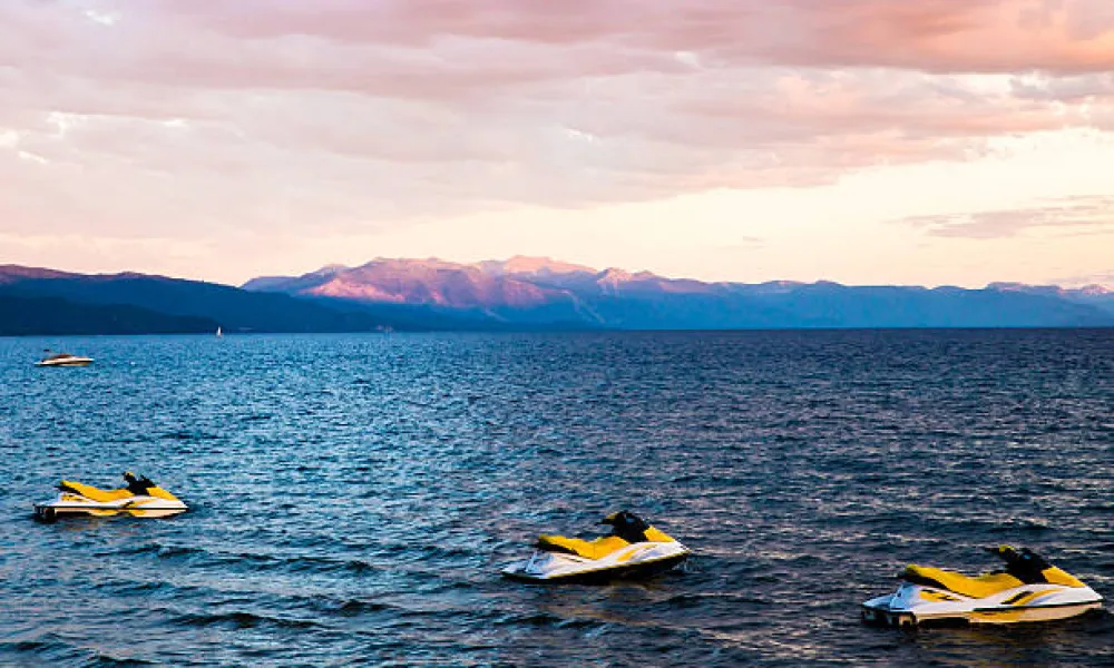 Three yellow and white jet skis floating on a lake with mountain range and pink sky at sunset.
