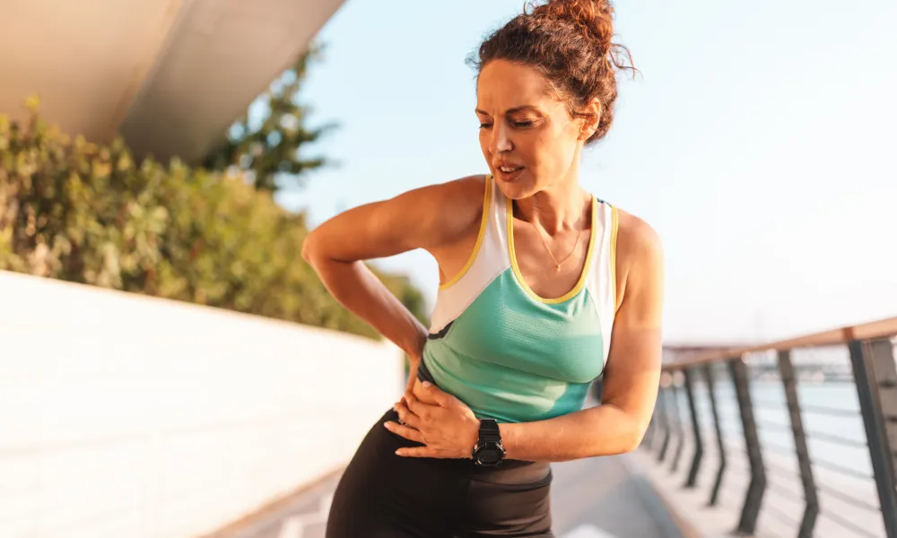 Woman in athletic wear clutching her side in pain while standing on outdoor pathway near water railing.