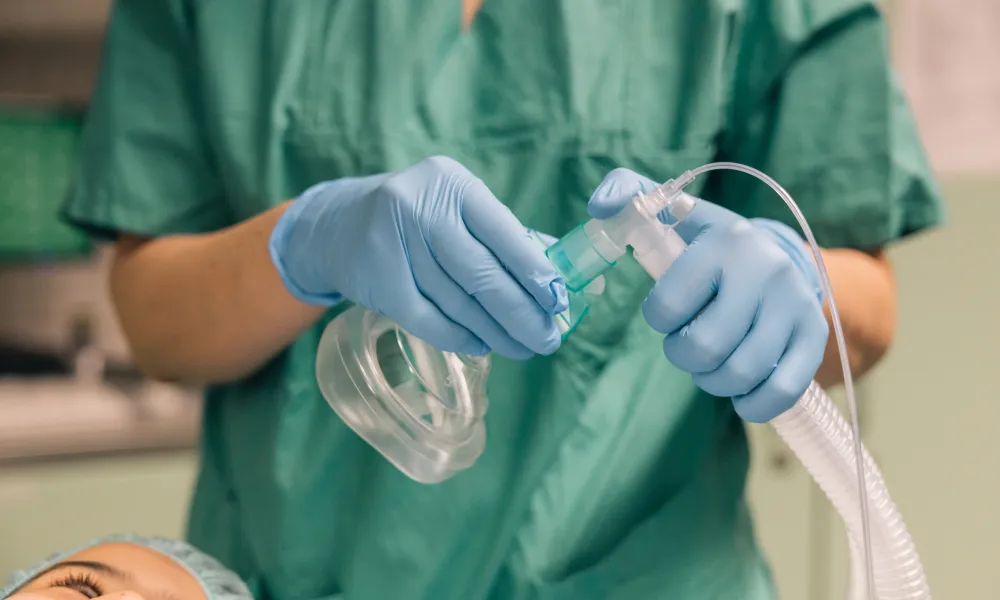 Medical professional in green scrubs wearing gloves adjusts anesthesia mask over patient's face in operating room.