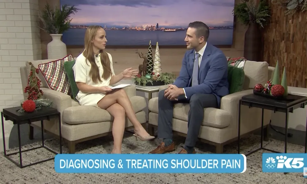 Two hosts discuss diagnosing and treating shoulder pain on a cozy TV set with holiday decor and cityscape backdrop.
