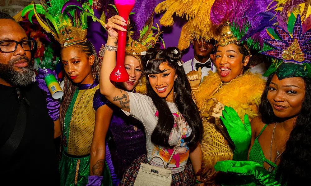 Group of people in colorful Mardi Gras costumes and feathers celebrating with drinks at a lively party.