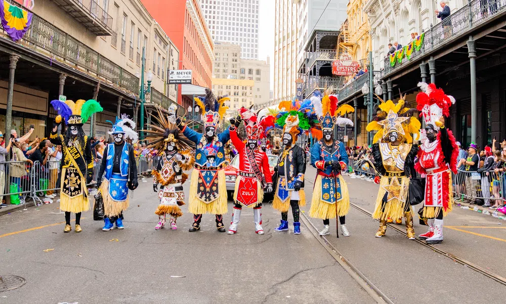 A colorful street parade with participants in feathered costumes and painted faces in an urban setting.