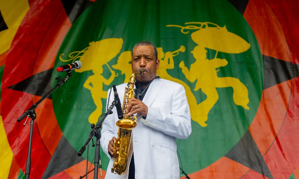 Saxophonist in white jacket performing on stage with colorful backdrop featuring silhouettes of musicians.