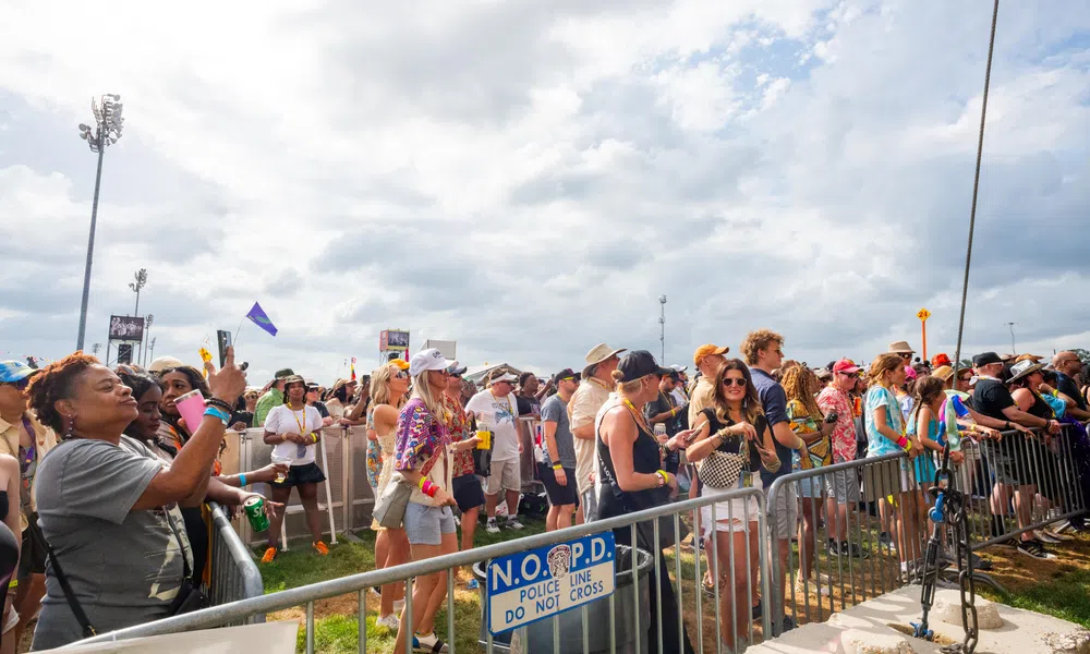 Diverse crowd enjoying an outdoor event behind police barriers under a cloudy sky with festival attire and accessories.