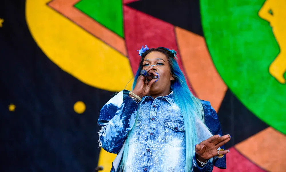 Female singer with blue ombre hair performing on stage with colorful abstract background art.