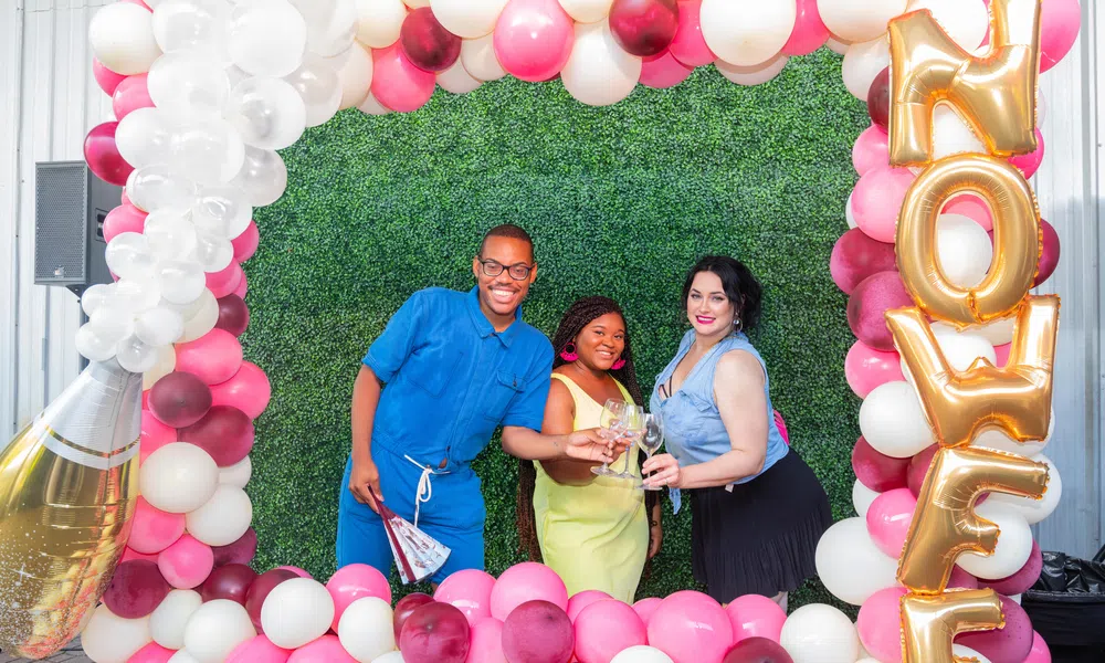 Three friends toasting drinks inside a pink and white balloon frame with gold 'NOWF' balloons at an outdoor party.