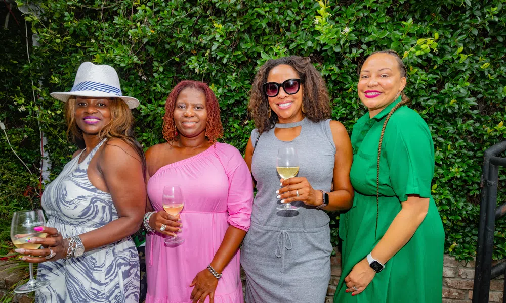 Four women dressed in colorful dresses holding wine glasses posing outdoors against green shrubbery.