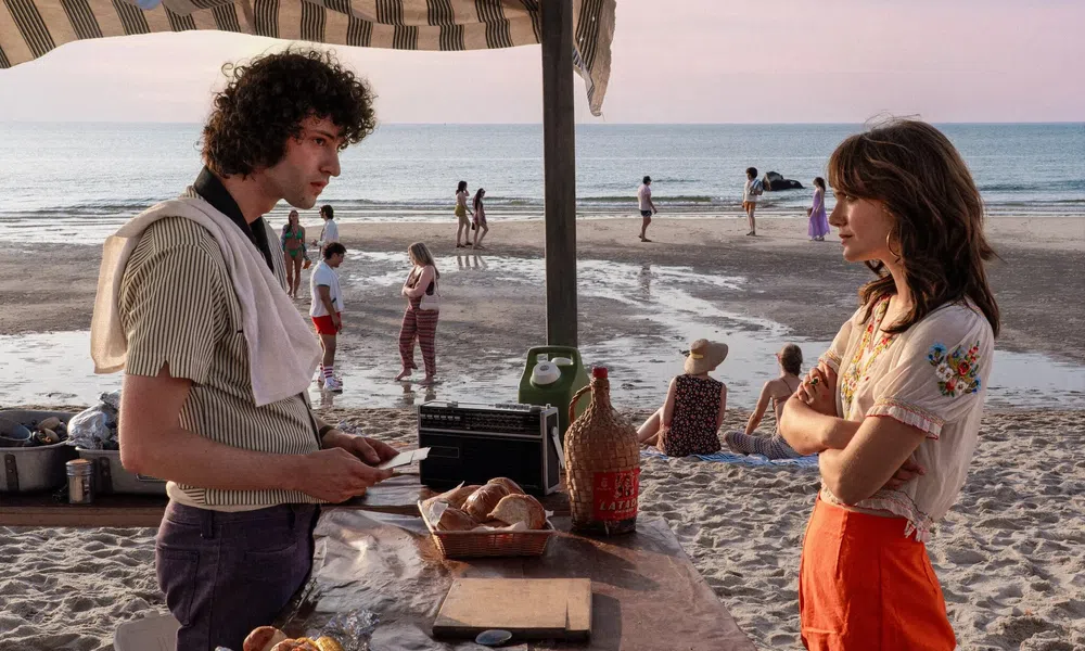 Young man and woman talking at a beachside stand with bread, drinks, and people walking along the shore at sunset.