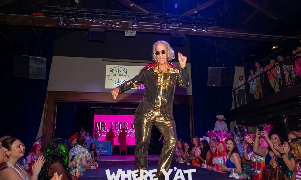 Older man in shiny gold outfit and platform shoes dancing on runway with cheering audience at event