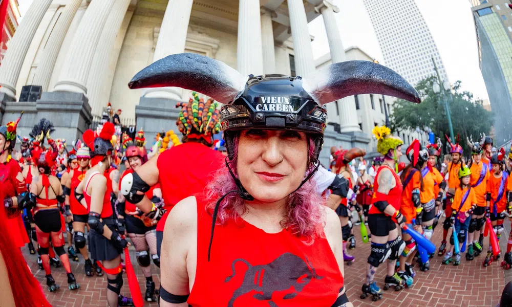 Person in red bull-themed helmet and shirt stands in front of a crowd in colorful costumes during outdoor event.