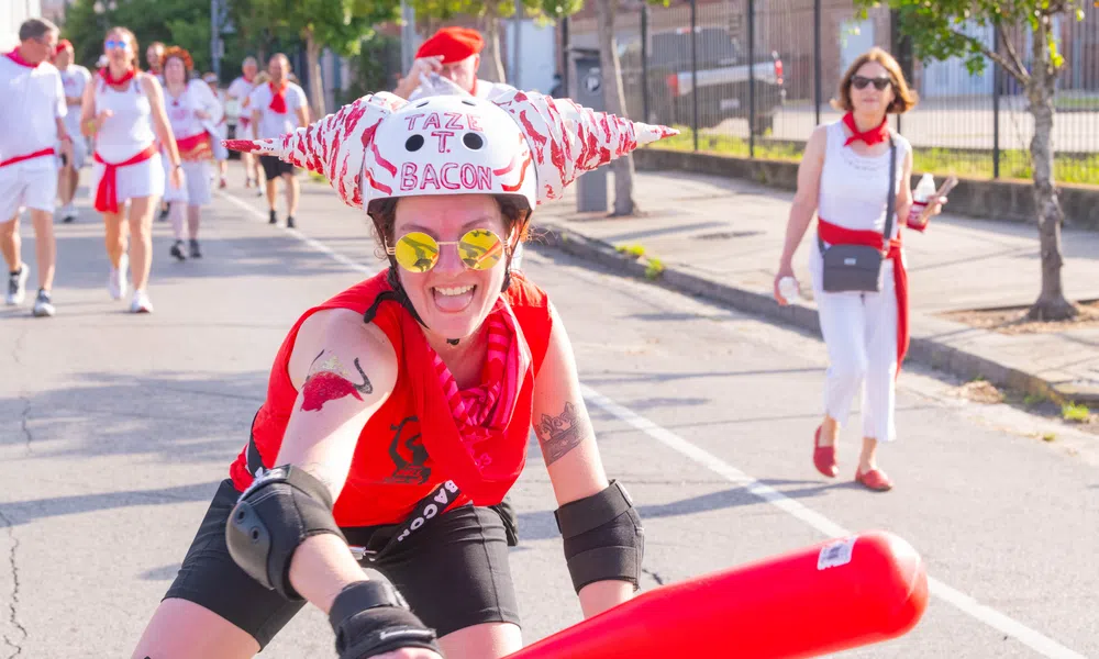 Person wearing a bacon-themed helmet and sunglasses holding a red bat during an outdoor event on a sunny day.