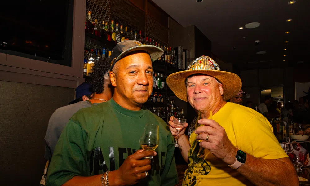 Two men enjoying drinks at a bar, one wearing a green shirt and hat, the other in a yellow shirt with a straw hat.