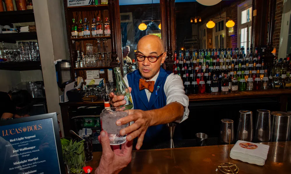 Bartender in a bow tie serving a cocktail at a wooden bar counter with bottles and glassware behind him