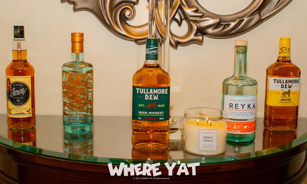 Five assorted liquor bottles and a lit French Vanilla candle displayed on a curved glass table with ornate metal wall decor.
