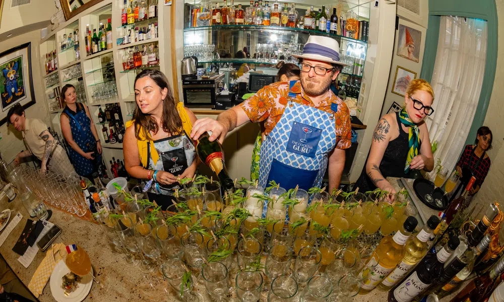 Bartenders preparing multiple cocktails with mint garnish in a busy bar setting with bottles and glasses around.