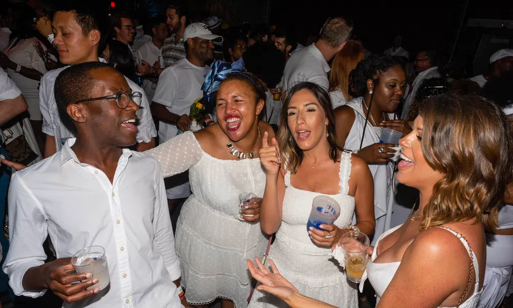 Group of friends in white outfits enjoying lively party with drinks and dancing at night event.