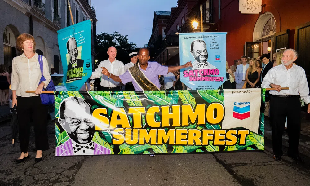 People holding a colorful Satchmo Summerfest banner during an evening parade in a city street.