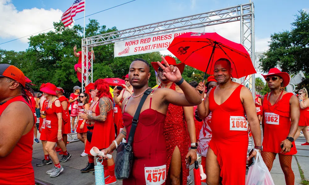 Participants dressed in red outfits posing at the NOH3 Red Dress Run start/finish line under a bright sky.